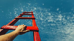 Eine Hand hält eine rote Leiter, die hinauf zum Himmel reicht.
