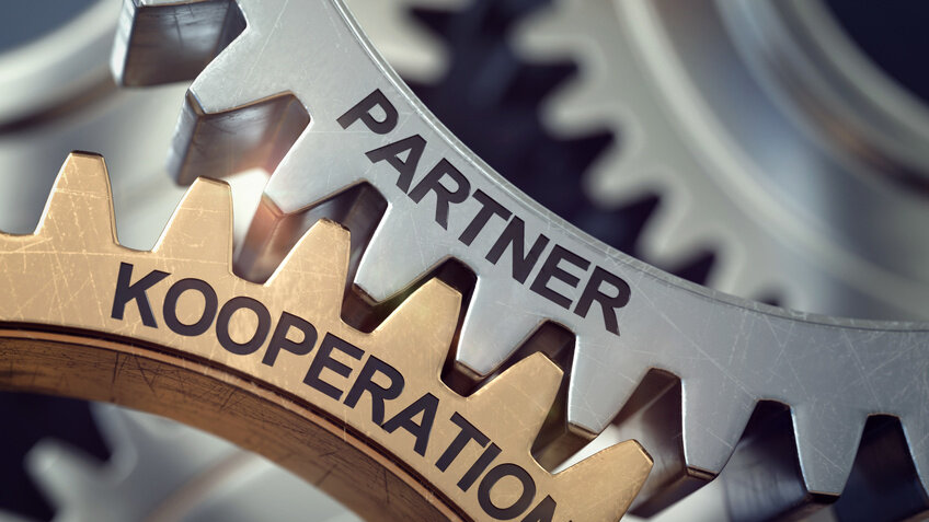 Zwei Zahnräder (silber und gold) greifen ineinander. Auf dem einen steht das Wort "Kooperation", auf dem anderen "Partner".