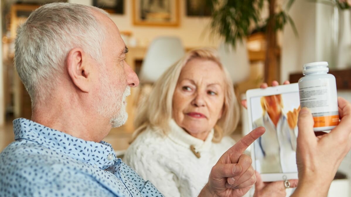 Alteres Ehepaar sitzt in einer Videosprechstunde am Tablet. Der ältere Mann zeigt der Ärztin im Bildschirm seine Medikamente.