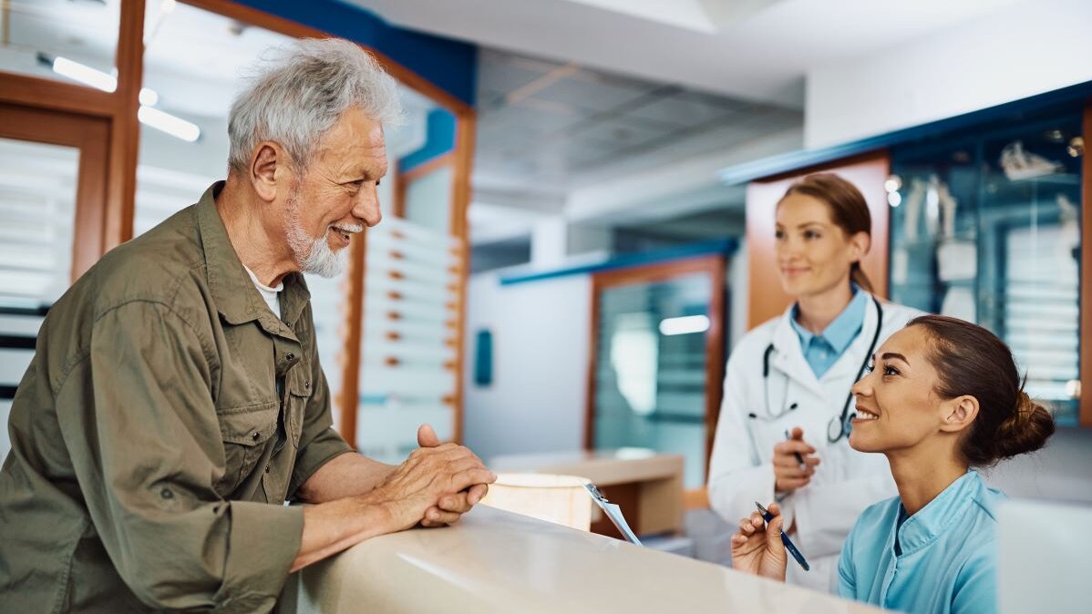 Ein älterer Herr steht an der Anmeldung in einer Arztpraxis und wird von einer Arzthelferin freundlich begrüßt.